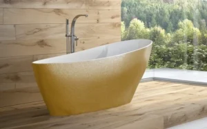 Złota wanna wolnostojąca Besco Keya Glam 165x70 w łazience z drewnianym wykończeniem i widokiem przez okno na las.