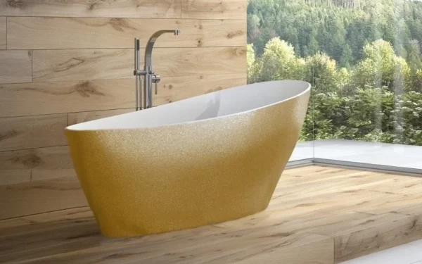 Złota wanna wolnostojąca Besco Keya Glam 165x70 w łazience z drewnianym wykończeniem i widokiem przez okno na las.