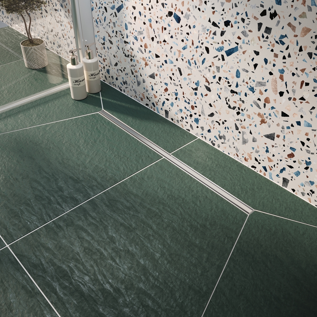 Vogi Ultra Slim Srebrny Srebrny, wąski odpływ liniowy VOGI Ultra Slim zamontowany przy ścianie z jasnego lastryko i beżowych płytek podłogowych.